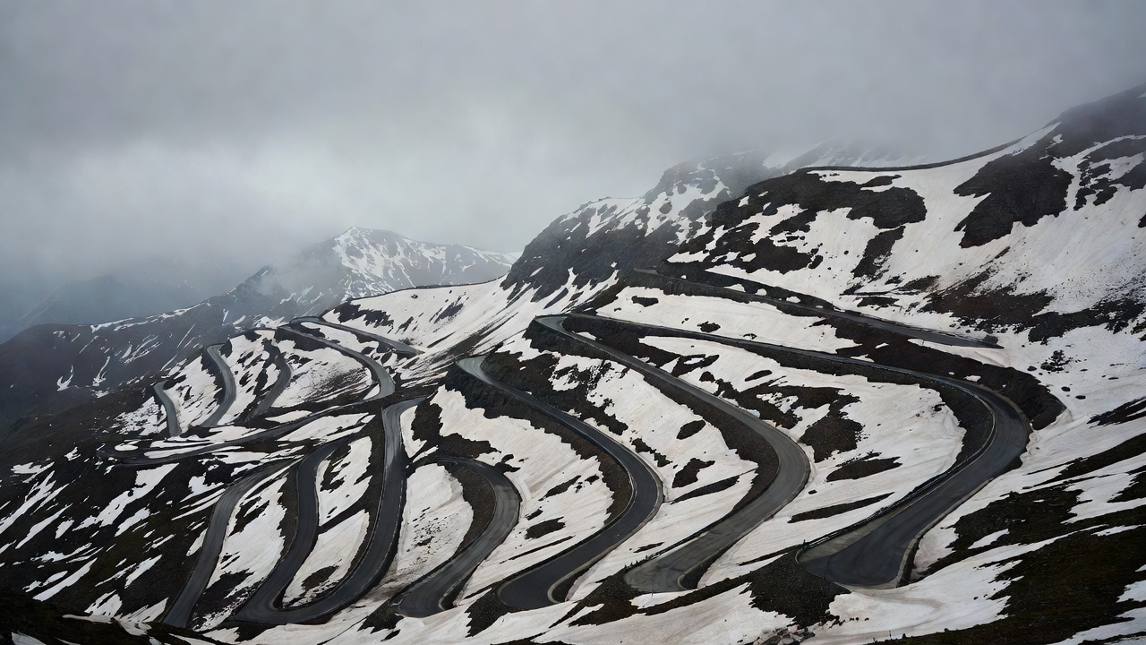 Stacked hairpin bends of the Stelvio Pass under low overcast cloud, snow patches on dark rock above the road
